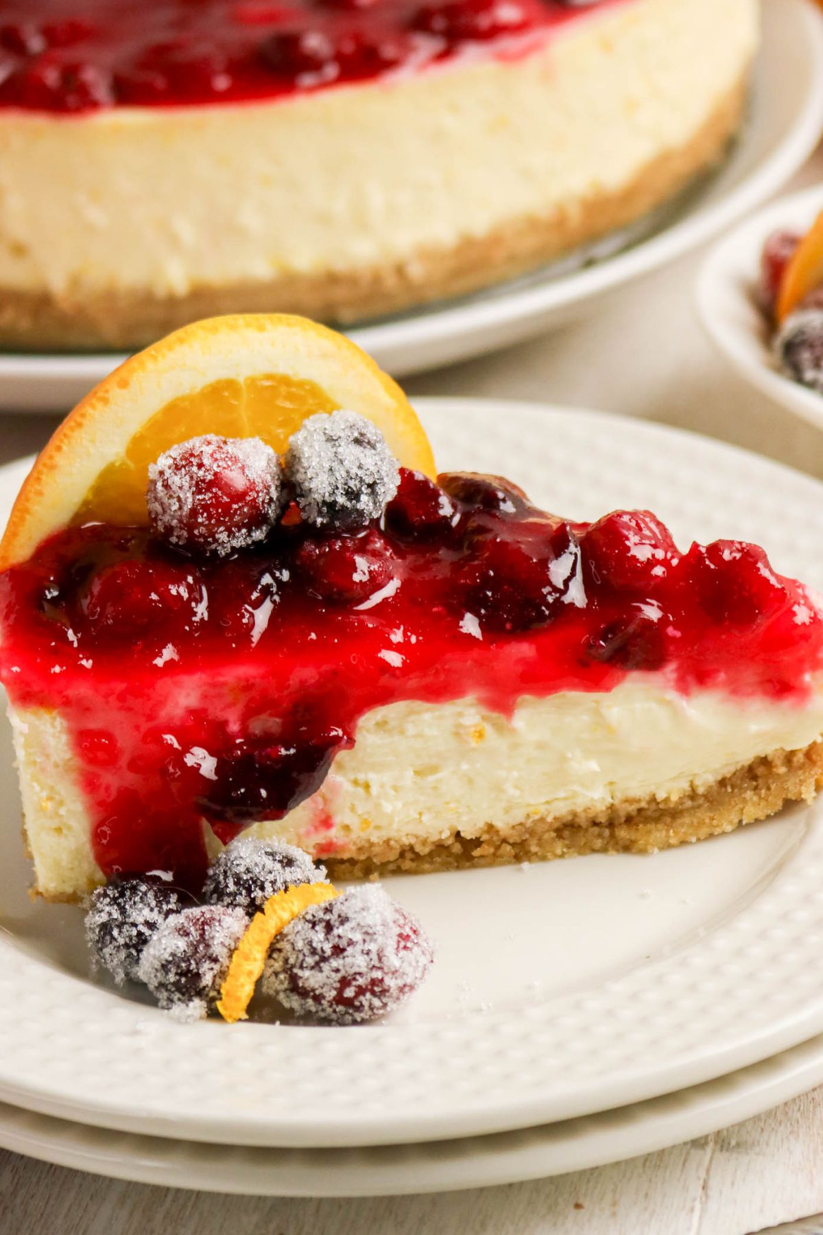 A slice of cheesecake topped with bright red berry sauce, sugared berries, and an orange slice, served on a white plate.