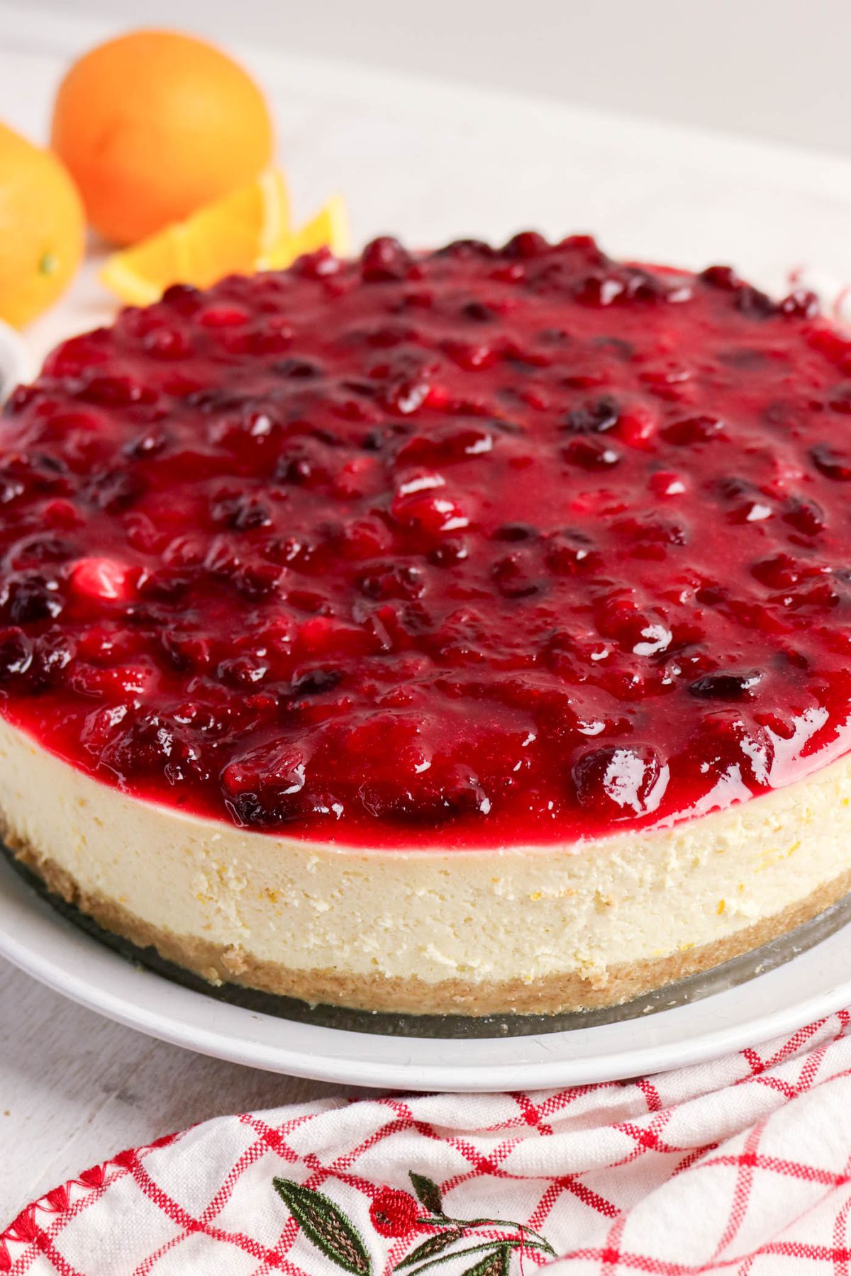 A cheesecake topped with a glossy mixed berry sauce sits on a white plate, with whole oranges in the background.
