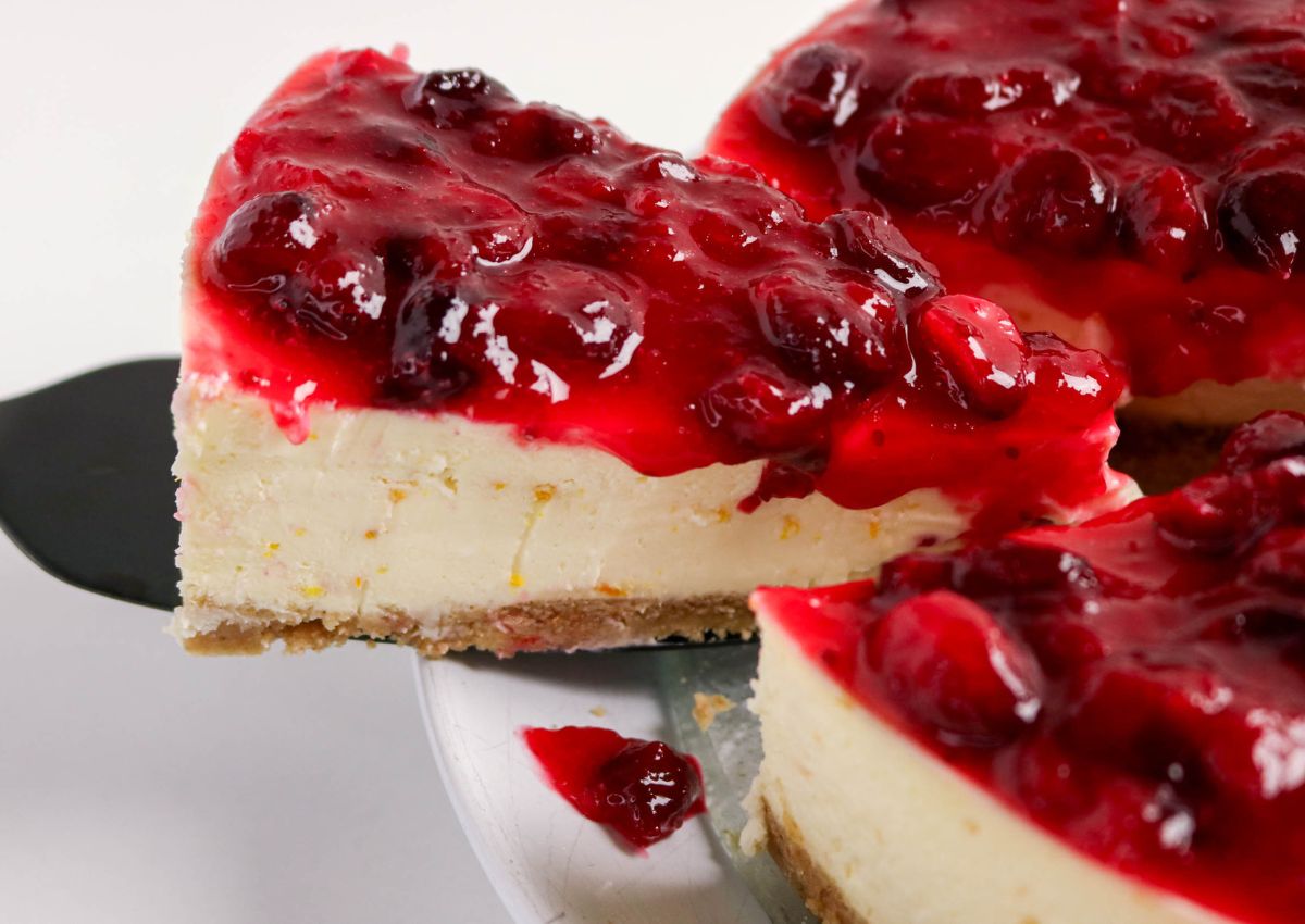 A slice of cheesecake with a graham cracker crust and a topping of glazed cherries is being served from a plate.