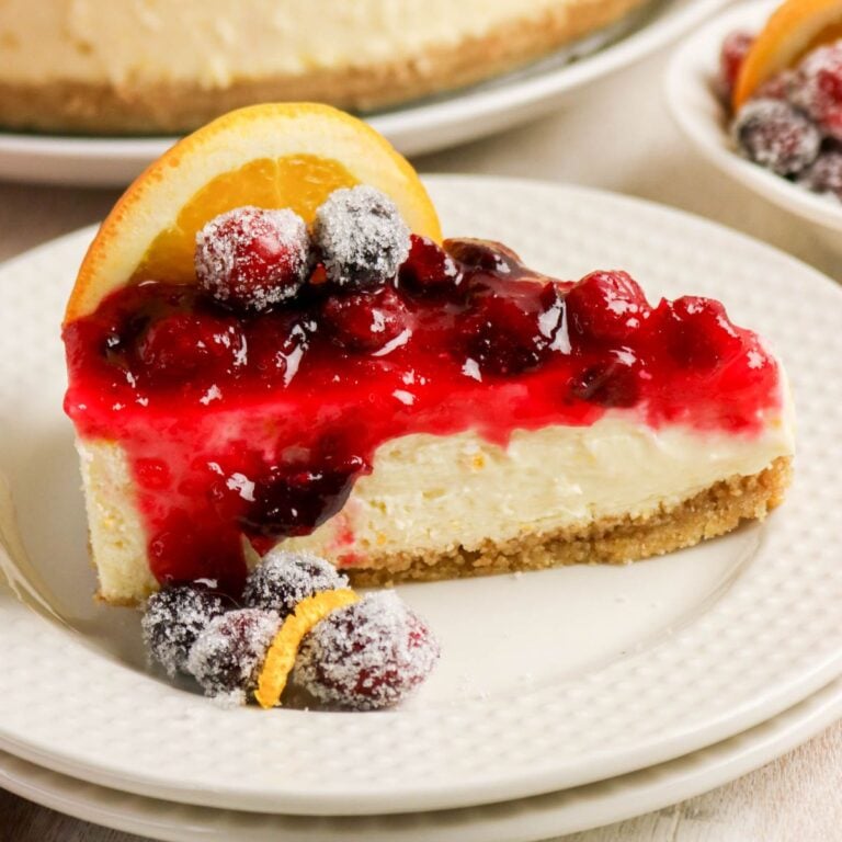 A slice of cheesecake with a graham cracker crust, topped with red berry sauce, sugared berries, and an orange slice, served on a white plate.