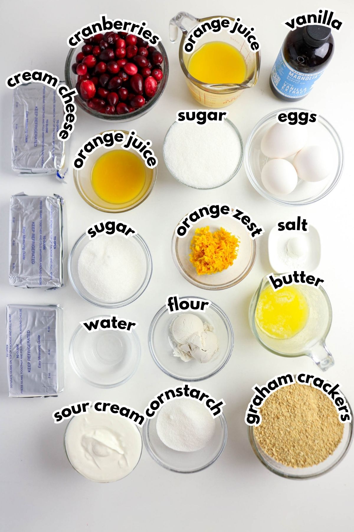 Top-down view of cheesecake ingredients in bowls and packages, including cream cheese, cranberries, orange juice, sugar, eggs, vanilla, sour cream, flour, cornstarch, salt, butter, and graham crackers.