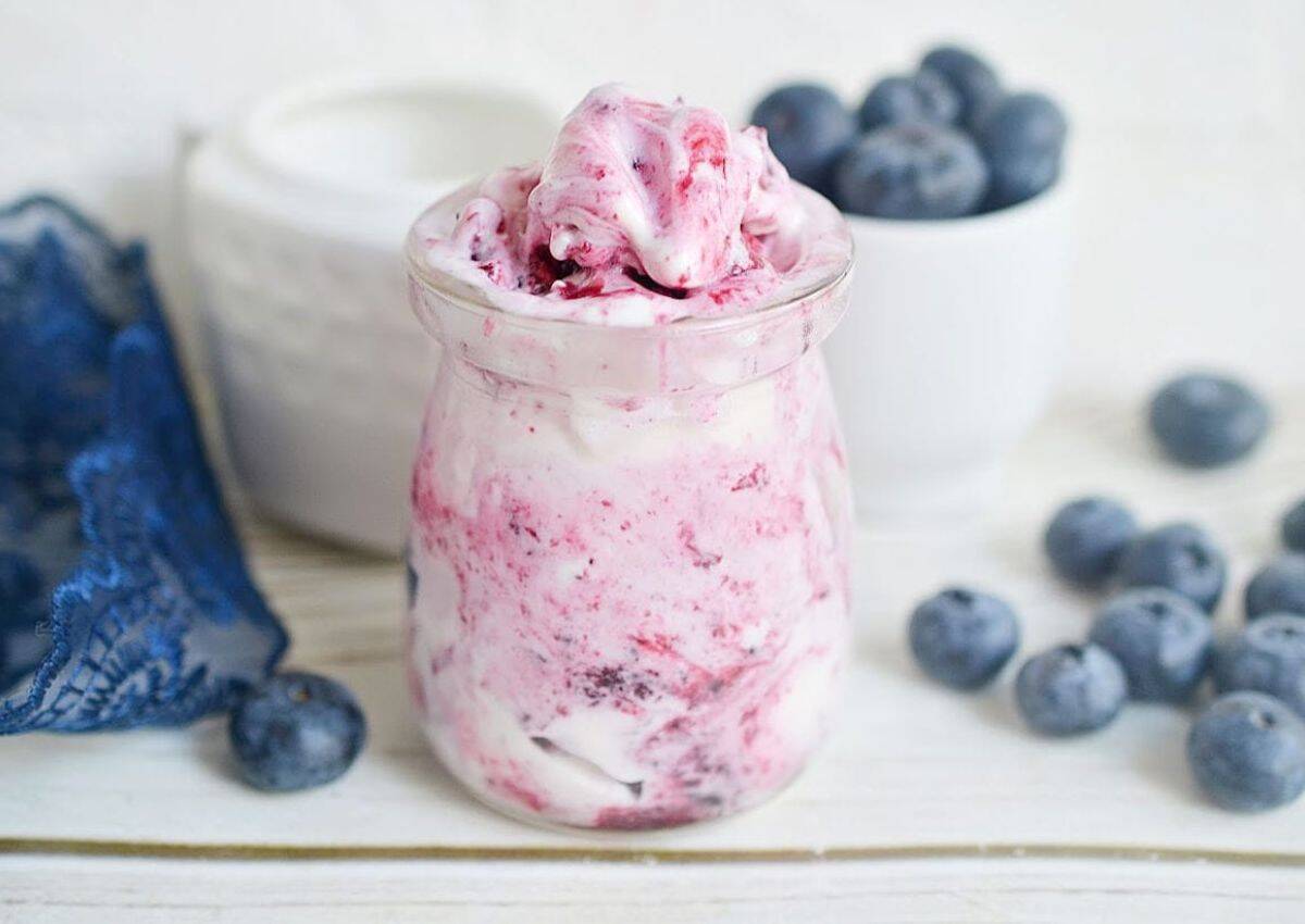 A glass jar of blueberry swirl yogurt sits on a white surface, surrounded by fresh blueberries and a small white bowl filled with more blueberries.