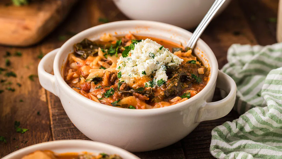A bowl of tomato-based soup with greens, rice, and a dollop of ricotta cheese on top, with a spoon in the bowl, on a wooden table.