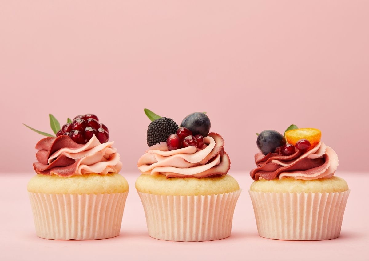 Three vanilla cupcakes with pink and red frosting are topped with berries and fruit, displayed in a row against a pale pink background.