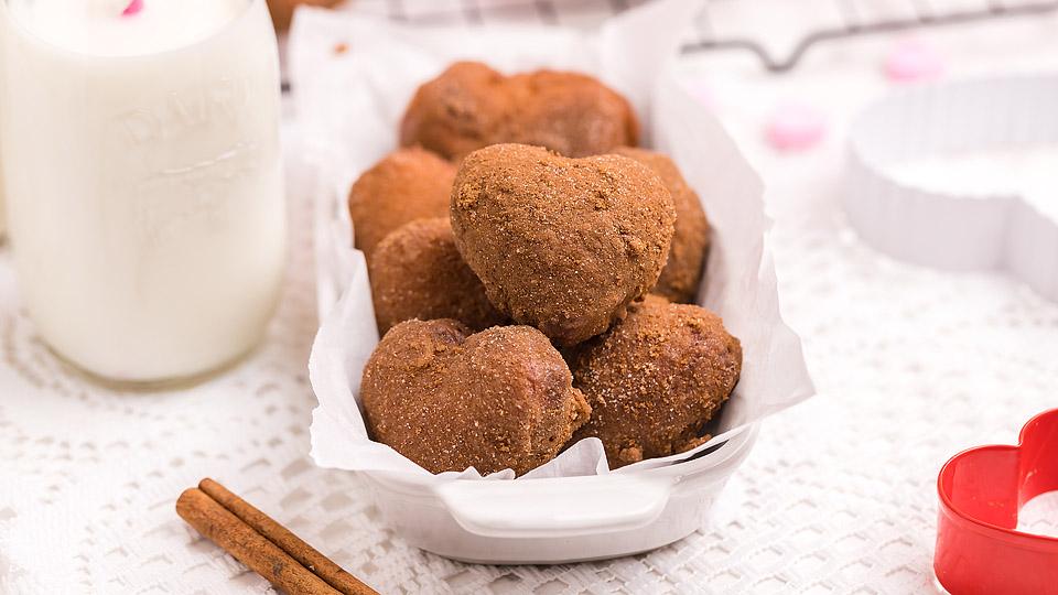 A white dish filled with heart-shaped, cinnamon-sugar coated pastries sits on a lace tablecloth next to a glass of milk and cinnamon sticks.