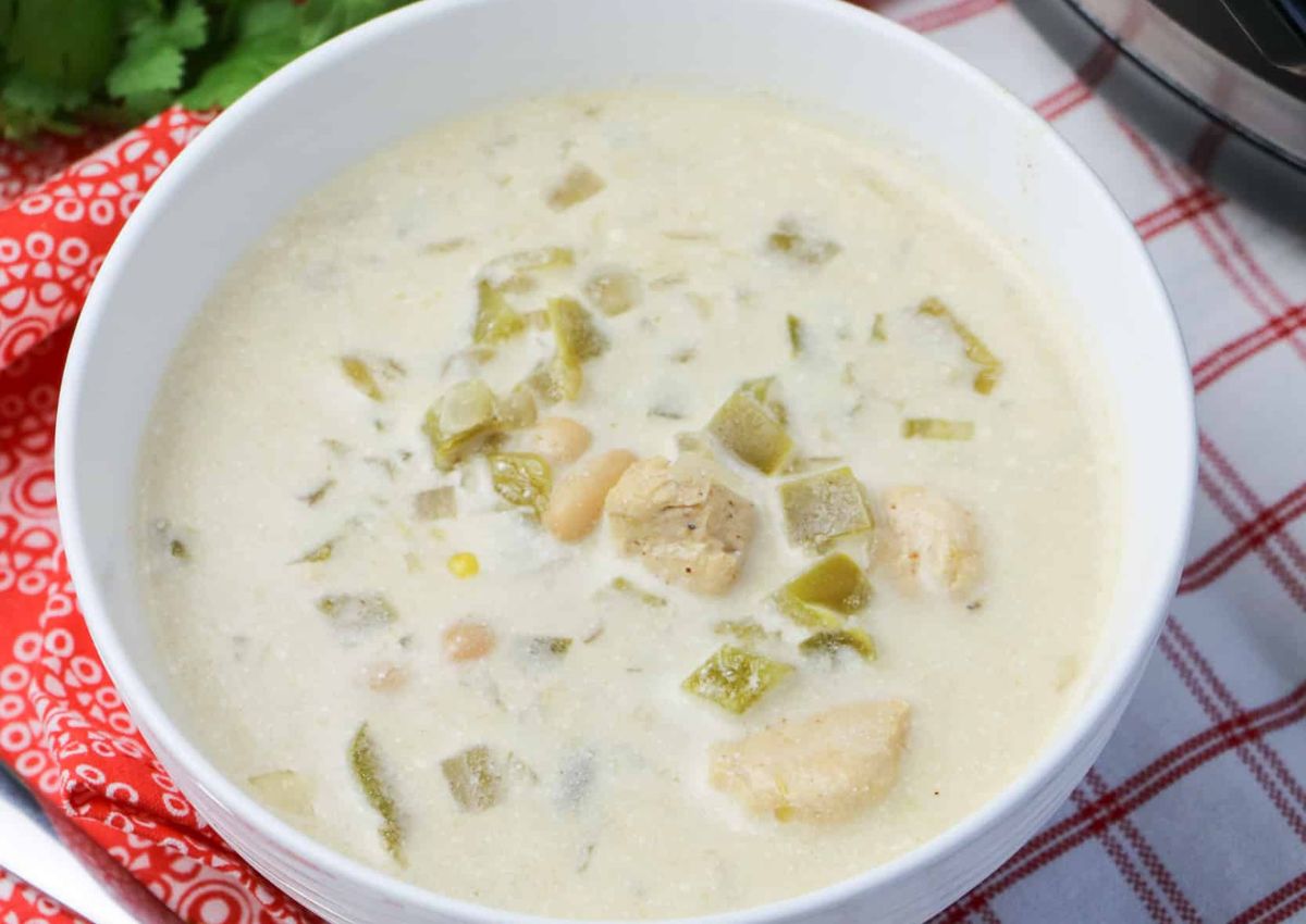 A bowl of creamy white chili with chunks of chicken and green chiles, placed on a red and white checkered cloth.