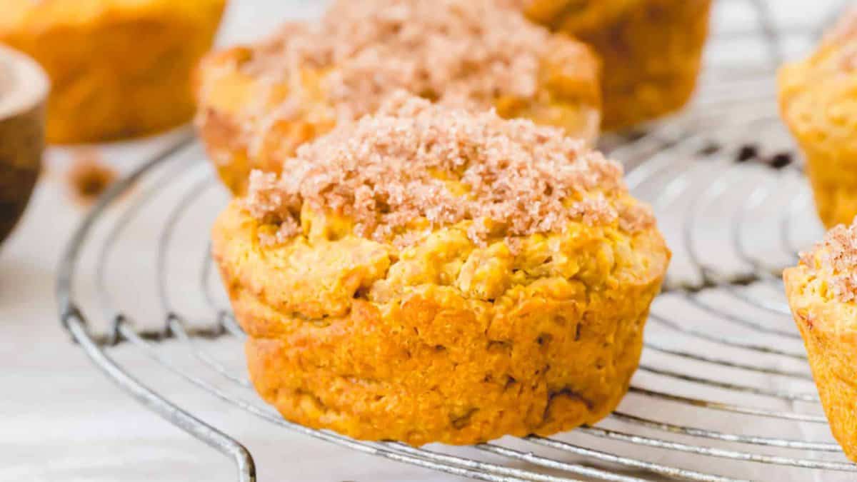 A close-up of a muffin with a crumbly topping, resting on a round metal cooling rack with other muffins in the background.