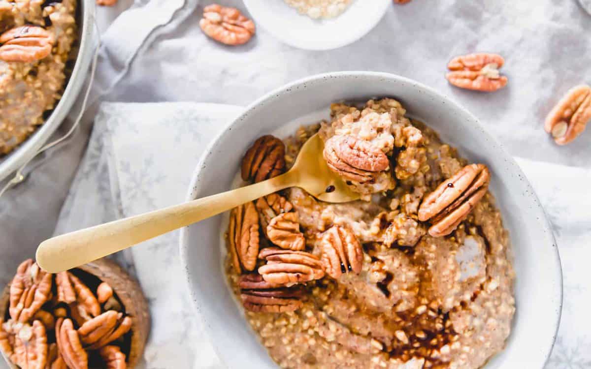 A bowl of oatmeal topped with whole pecans and a drizzle of syrup, with a gold-colored spoon resting inside.