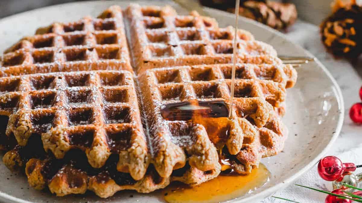A stack of waffles on a plate with syrup being poured over them, dusted with powdered sugar.