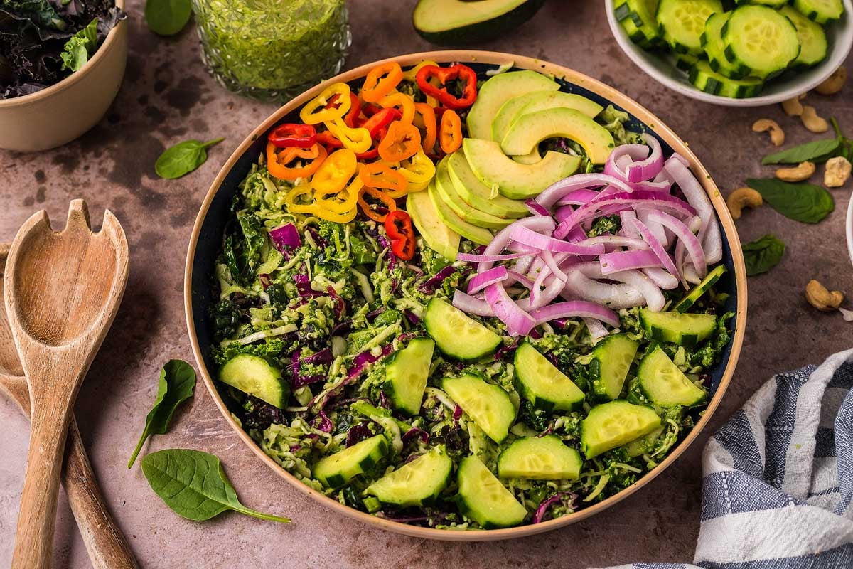 A bowl of salad with sliced cucumber, red onion, avocado, bell peppers, and shredded greens on a table with salad utensils and extra vegetables.