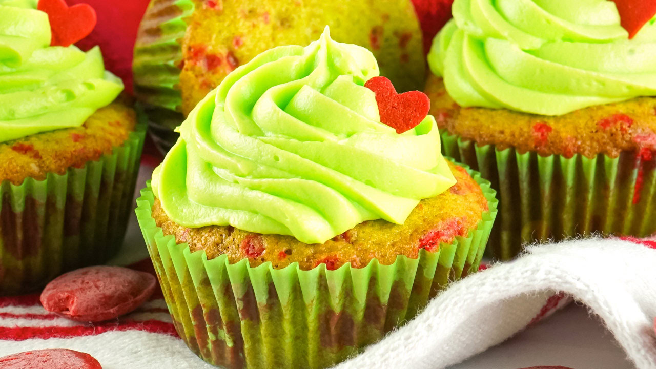 Cupcakes with green frosting and red heart decorations in green cupcake liners, arranged on a surface with a white and red striped cloth.