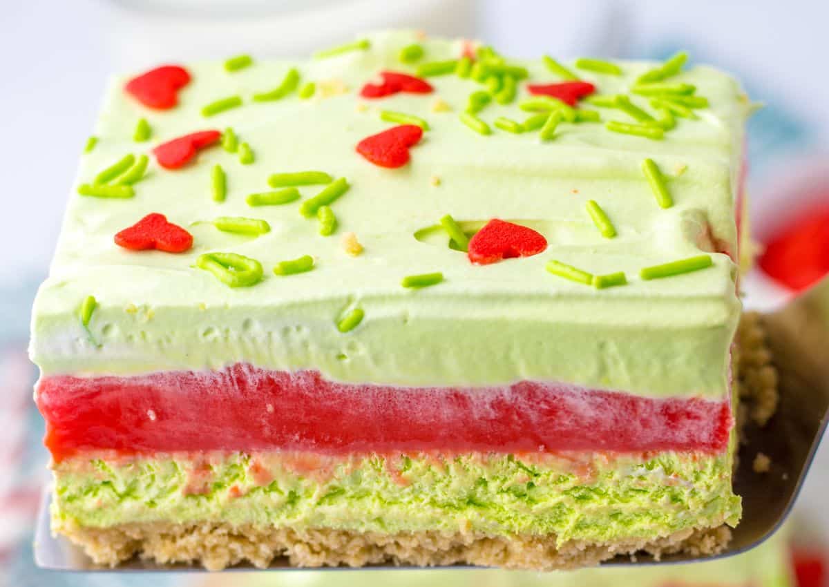 A close-up of a dessert bar with a crumbly base, green and red layers, topped with green sprinkles and red heart-shaped decorations.