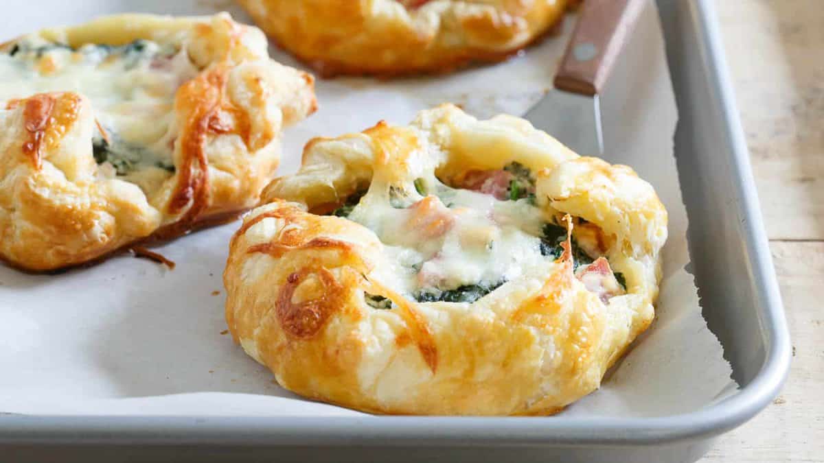 A golden-brown puff pastry tart filled with melted cheese, spinach, and diced ham sits on a parchment-lined baking tray with a serving knife nearby.