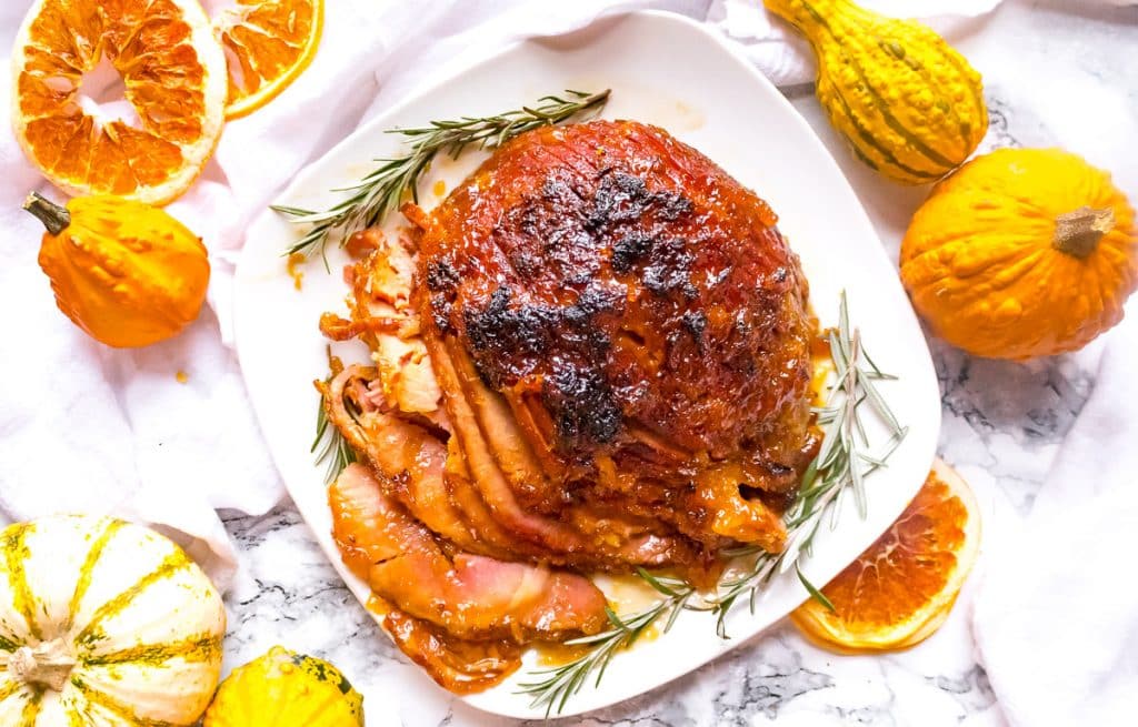A glazed ham garnished with rosemary sits on a white plate, surrounded by dried orange slices and decorative gourds on a marble surface.