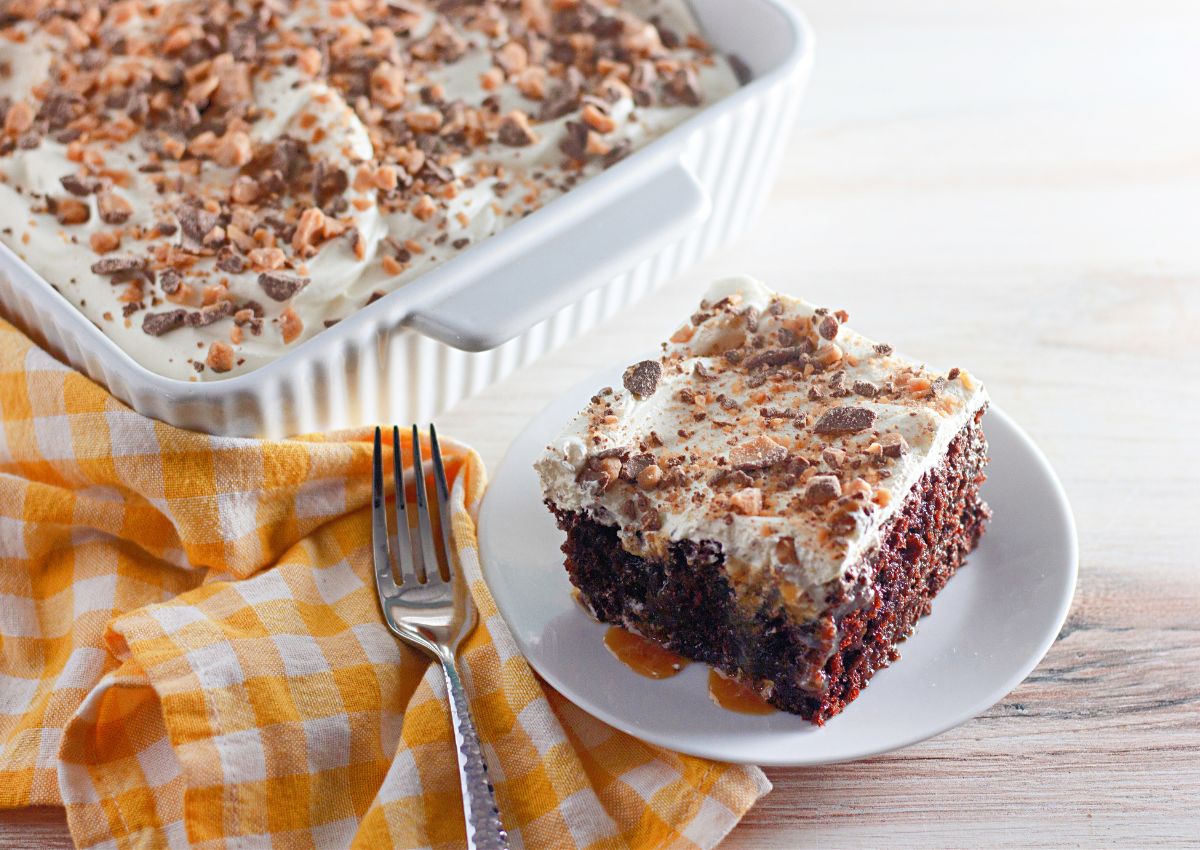 A slice of chocolate cake with whipped topping and crushed toffee sits on a plate next to a baking dish with more cake, a fork, and a yellow checked cloth.