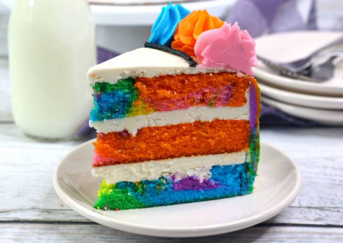 A slice of rainbow layered cake with white frosting and colorful decorative icing on top, served on a white plate.