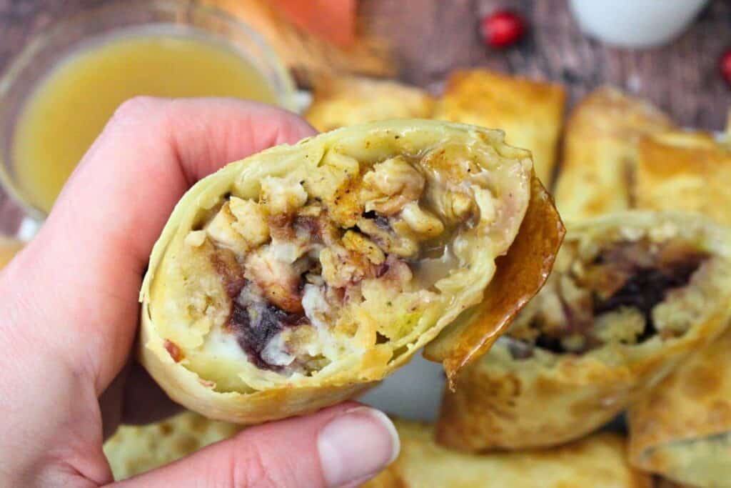 A hand holds a cut-open egg roll filled with stuffing, turkey, and other Thanksgiving ingredients; whole egg rolls and a dipping sauce are in the background.