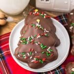 Three chocolate-covered cookies shaped like Christmas trees, topped with red, green, and white sprinkles, on a white plate next to a red plaid cloth.
