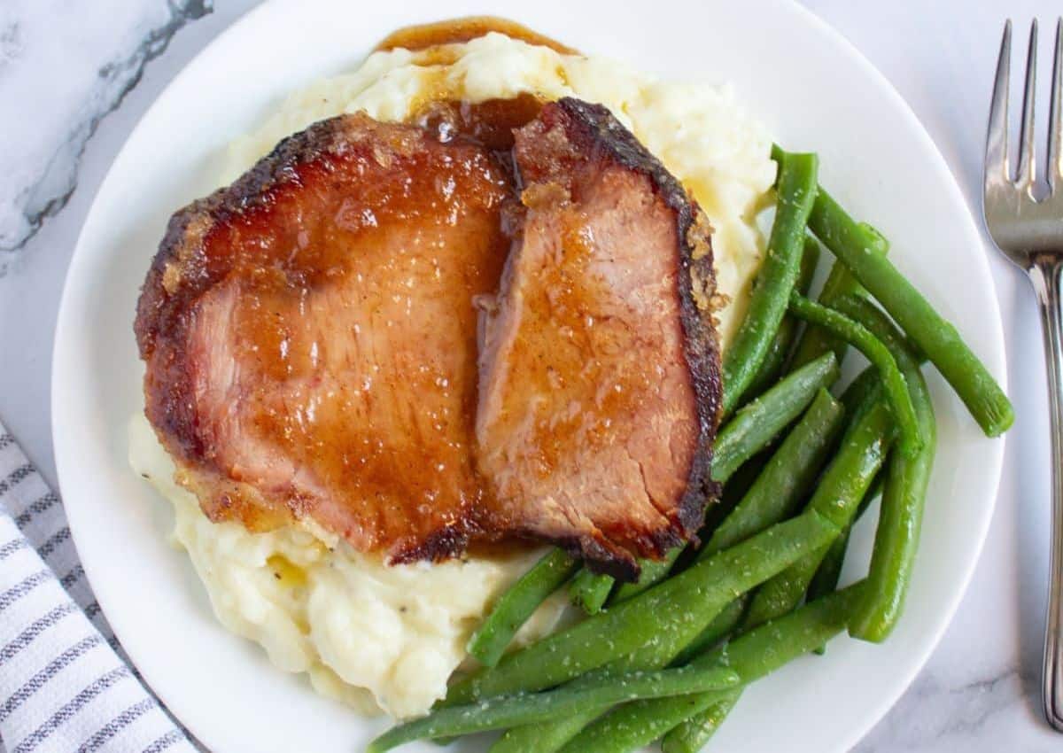 A plate with two slices of glazed pork roast on mashed potatoes, served with green beans. A fork is placed on the right side of the plate.