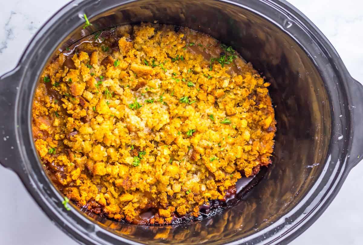 A slow cooker filled with baked stuffing topped with browned breadcrumbs and garnished with herbs.