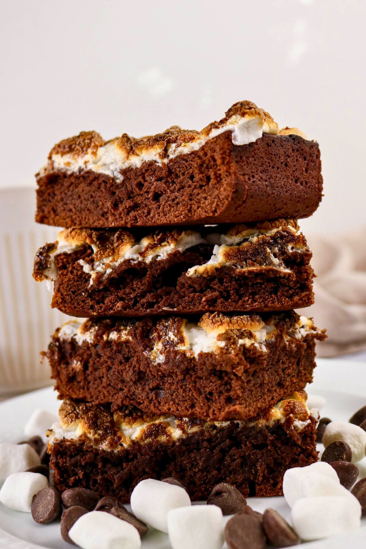 A stack of four chocolate brownies with melted marshmallows on top, surrounded by chocolate chips and mini marshmallows.