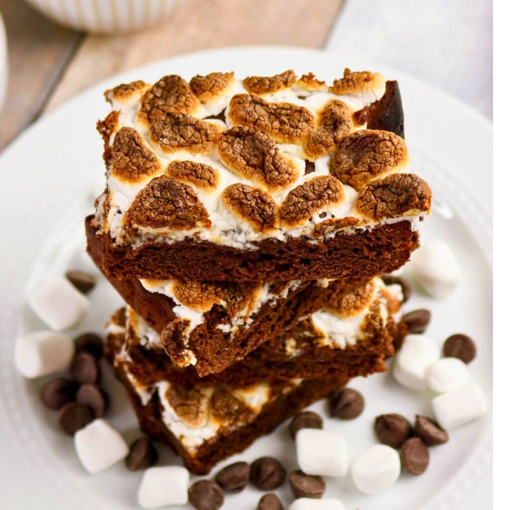 Three chocolate brownies topped with toasted marshmallows are stacked on a white plate, surrounded by chocolate chips and mini marshmallows.