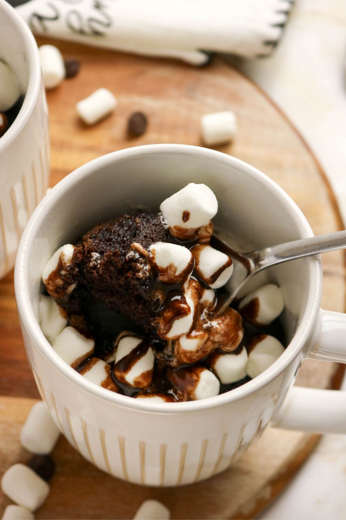 A white mug filled with chocolate mug cake, mini marshmallows, and chocolate syrup, with a spoon inside, sitting on a wooden surface.