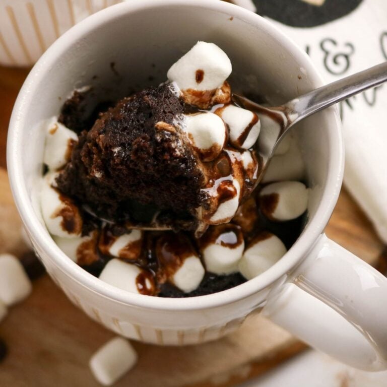 A mug filled with chocolate cake topped with mini marshmallows and chocolate drizzle, with a spoon scooping out a portion.