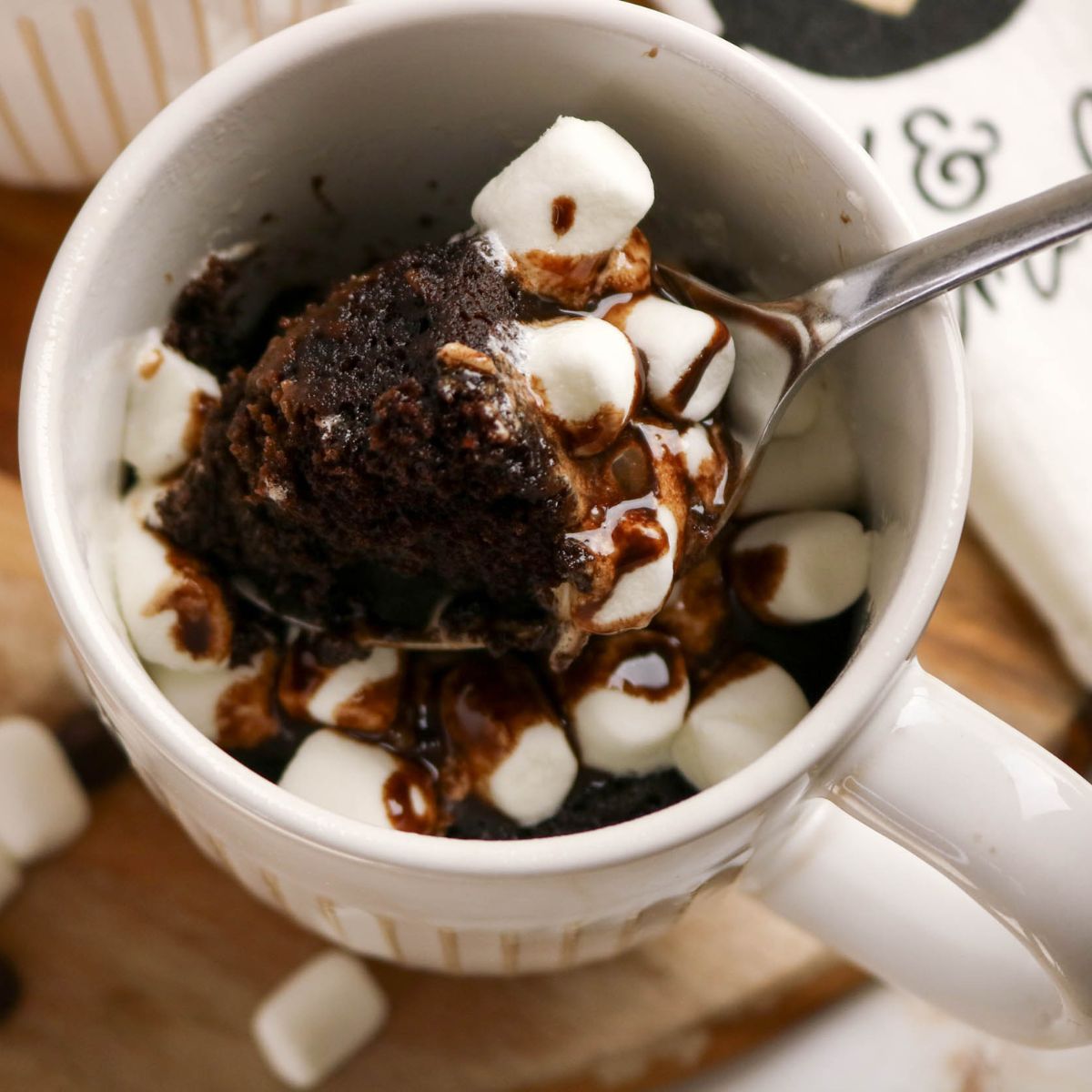 A mug filled with chocolate cake topped with mini marshmallows and chocolate drizzle, with a spoon scooping out a portion.