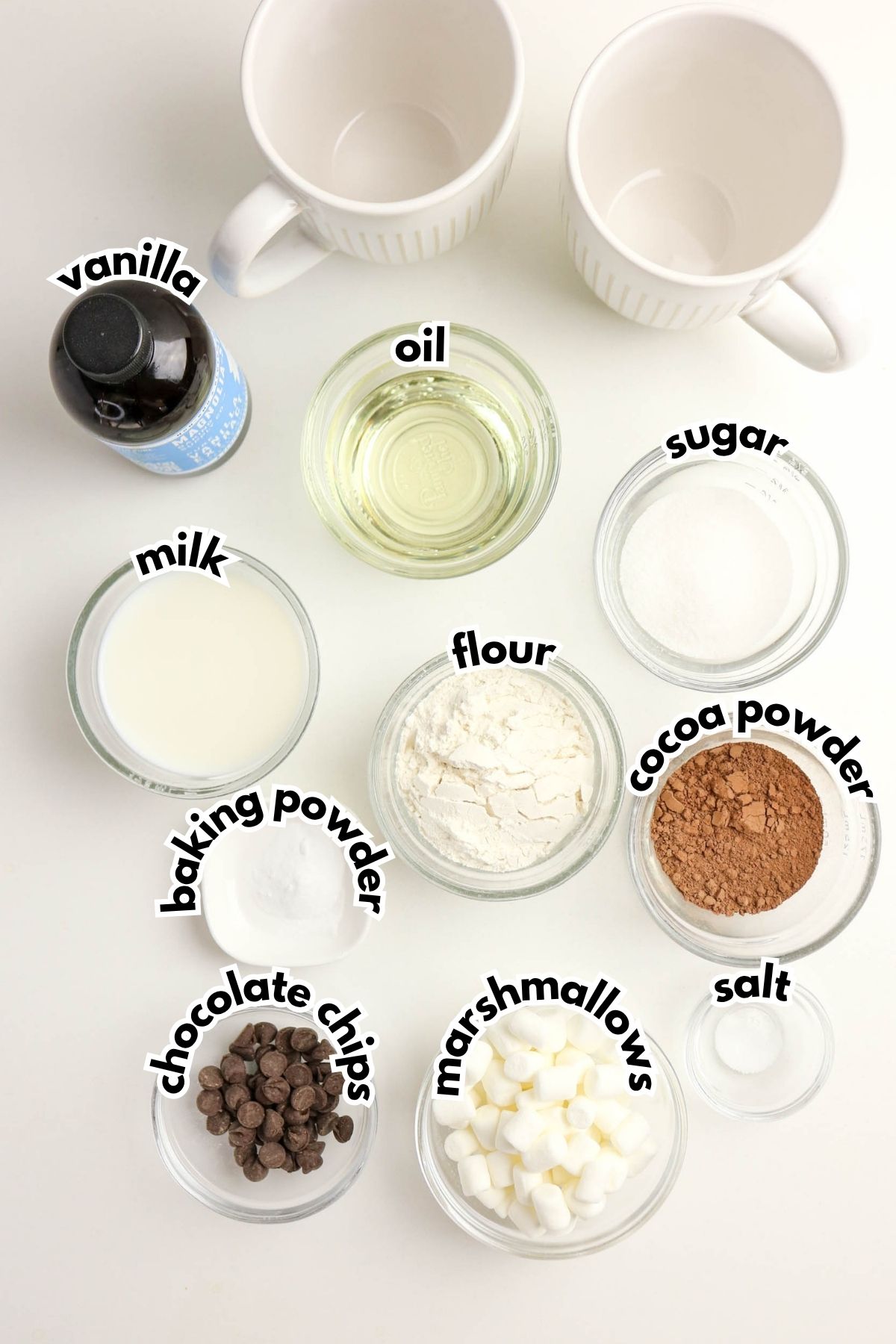 Two white mugs and labeled bowls containing vanilla, oil, sugar, milk, flour, baking powder, cocoa powder, salt, chocolate chips, and marshmallows on a white surface.