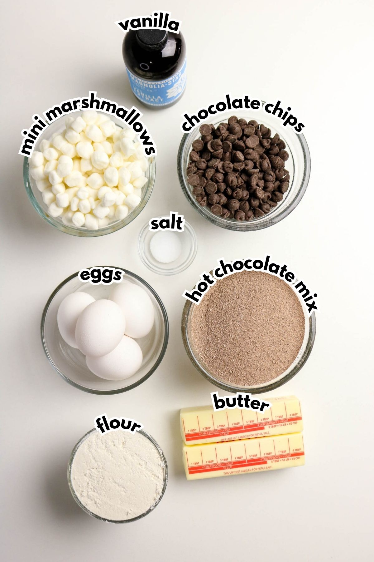 Bowls and containers holding mini marshmallows, chocolate chips, hot chocolate mix, flour, eggs, salt, vanilla extract, and sticks of butter are arranged on a white surface.