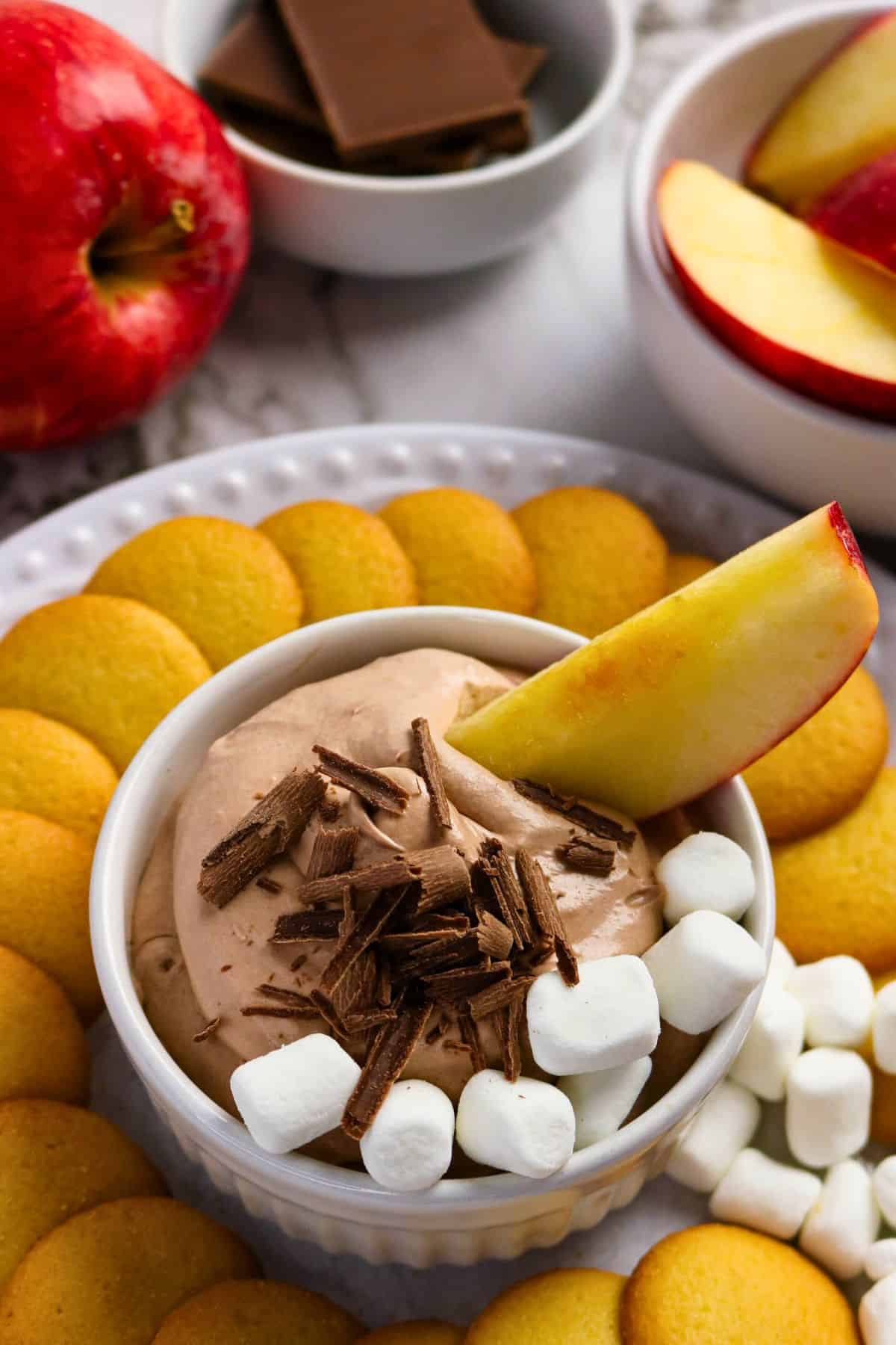 A bowl of chocolate dip topped with chocolate shavings, mini marshmallows, and an apple slice, surrounded by vanilla wafer cookies, apple slices, and chocolate squares.