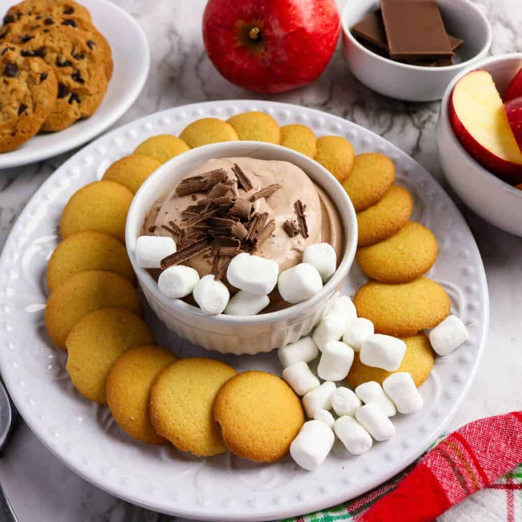 A plate with vanilla wafers, a bowl of chocolate dip topped with marshmallows and chocolate shavings, cookies, apple slices, and chocolate pieces on the side.