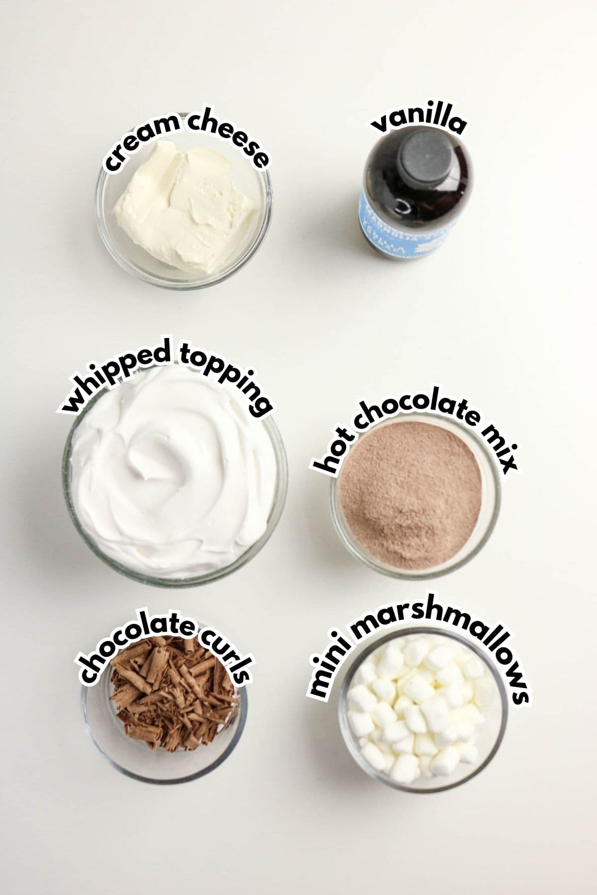 Six bowls on a white surface containing cream cheese, vanilla, whipped topping, hot chocolate mix, chocolate curls, and mini marshmallows, each labeled with text.