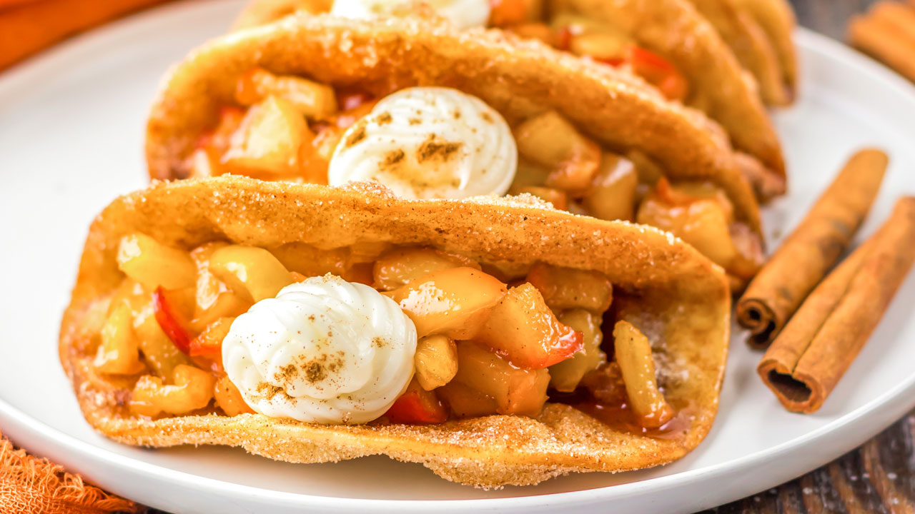 Three dessert tacos filled with chopped fruit and topped with whipped cream are arranged on a white plate next to two cinnamon sticks.