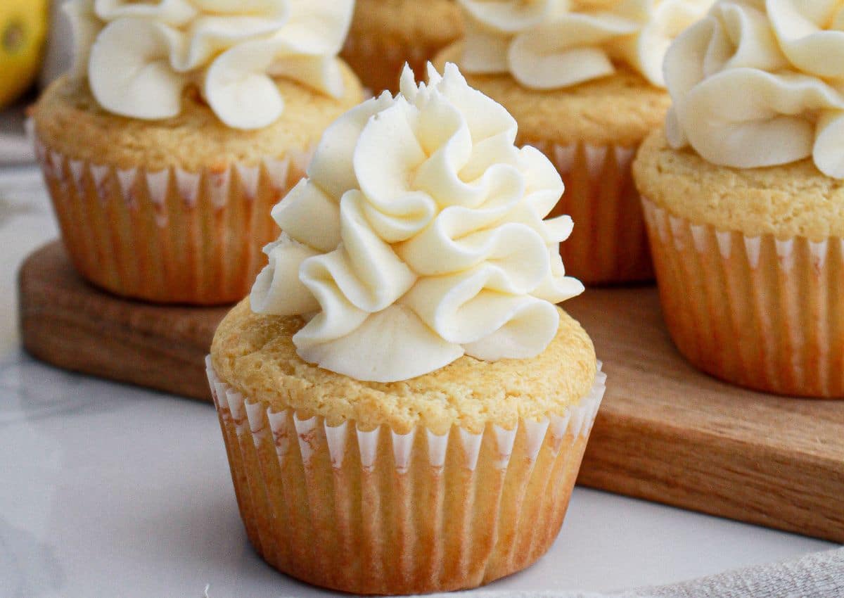 Vanilla cupcakes with swirled white buttercream frosting are arranged on a wooden board and a marble surface.