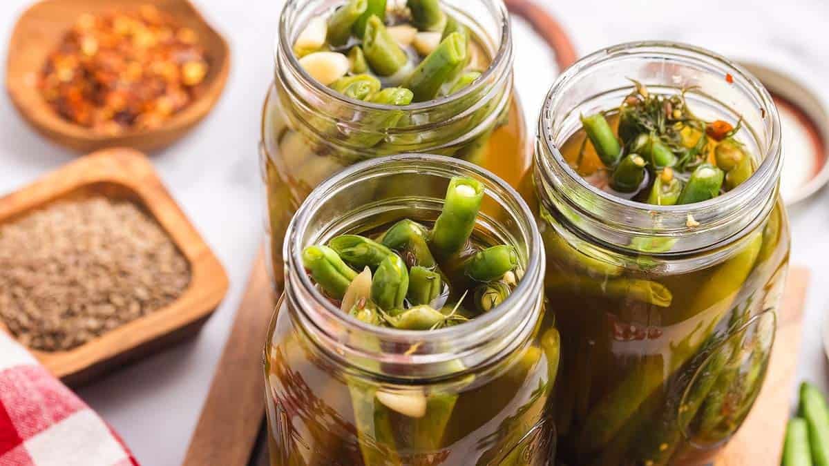 Three glass jars filled with green beans, herbs, and garlic in brine sit on a table, with containers of spices in the background.