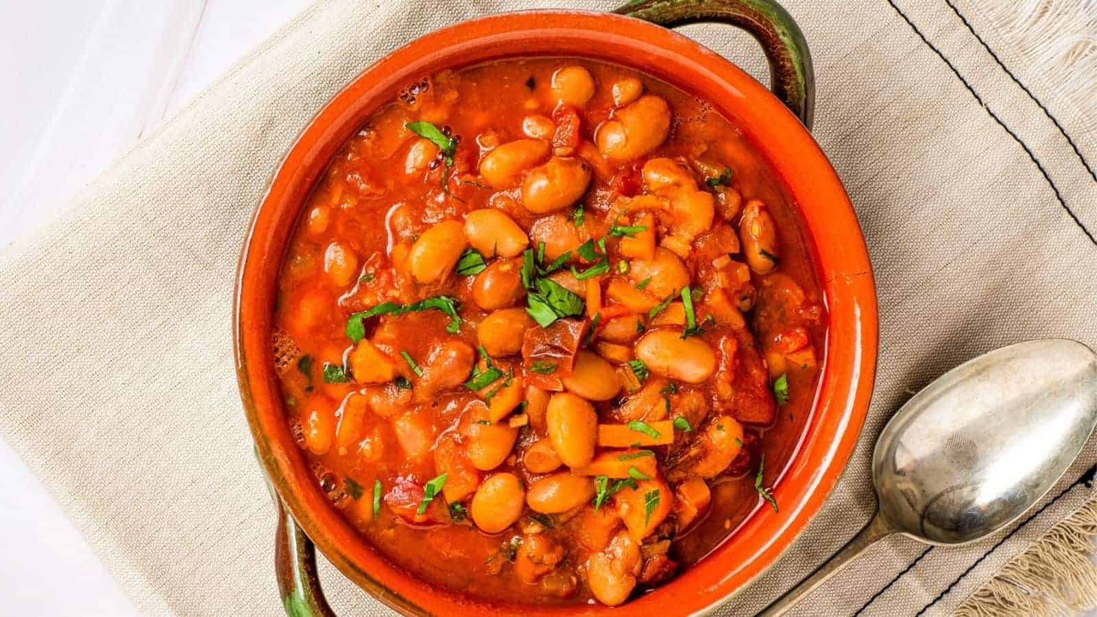 A bowl of baked beans in tomato sauce, garnished with chopped herbs, placed on a beige cloth next to a metal spoon.