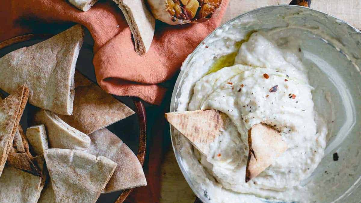 A bowl of creamy dip garnished with olive oil and spices, served with pita bread pieces on the side.