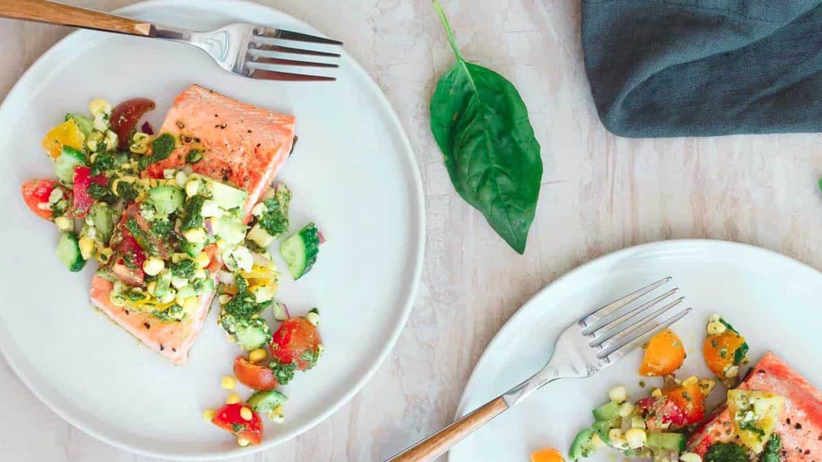 Two white plates with salmon fillets topped with a colorful vegetable salsa, each with a fork. A basil leaf and a dark napkin are on the table.
