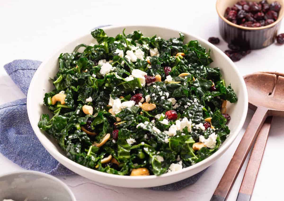 A bowl of kale salad with feta cheese, dried cranberries, and cashews on a white table, next to a wooden spoon and a small dish of cranberries.