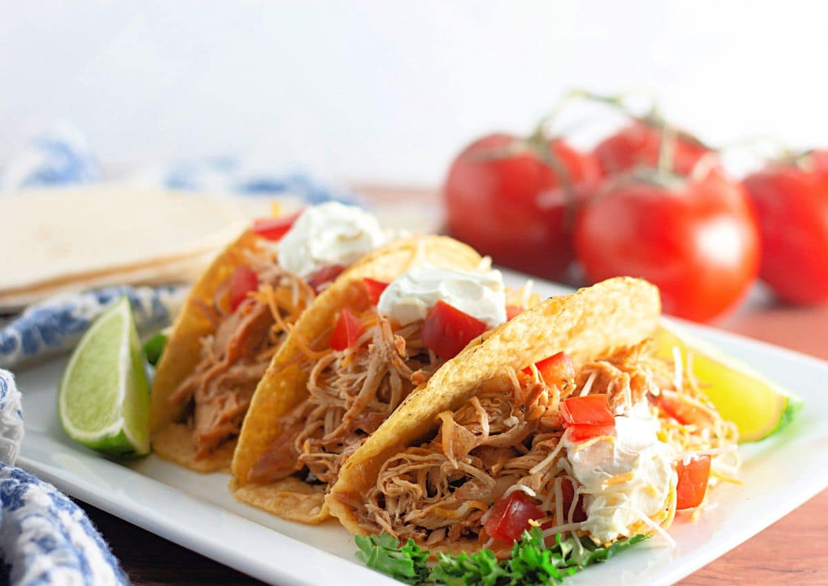 Three shredded chicken tacos with diced tomatoes, shredded cheese, and sour cream on a white plate, garnished with lime wedges and parsley, with tomatoes in the background.