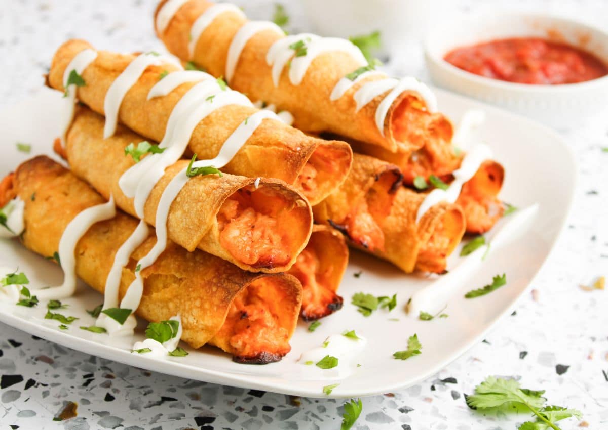 A plate of chicken taquitos drizzled with sour cream and garnished with chopped cilantro, with a bowl of salsa in the background.