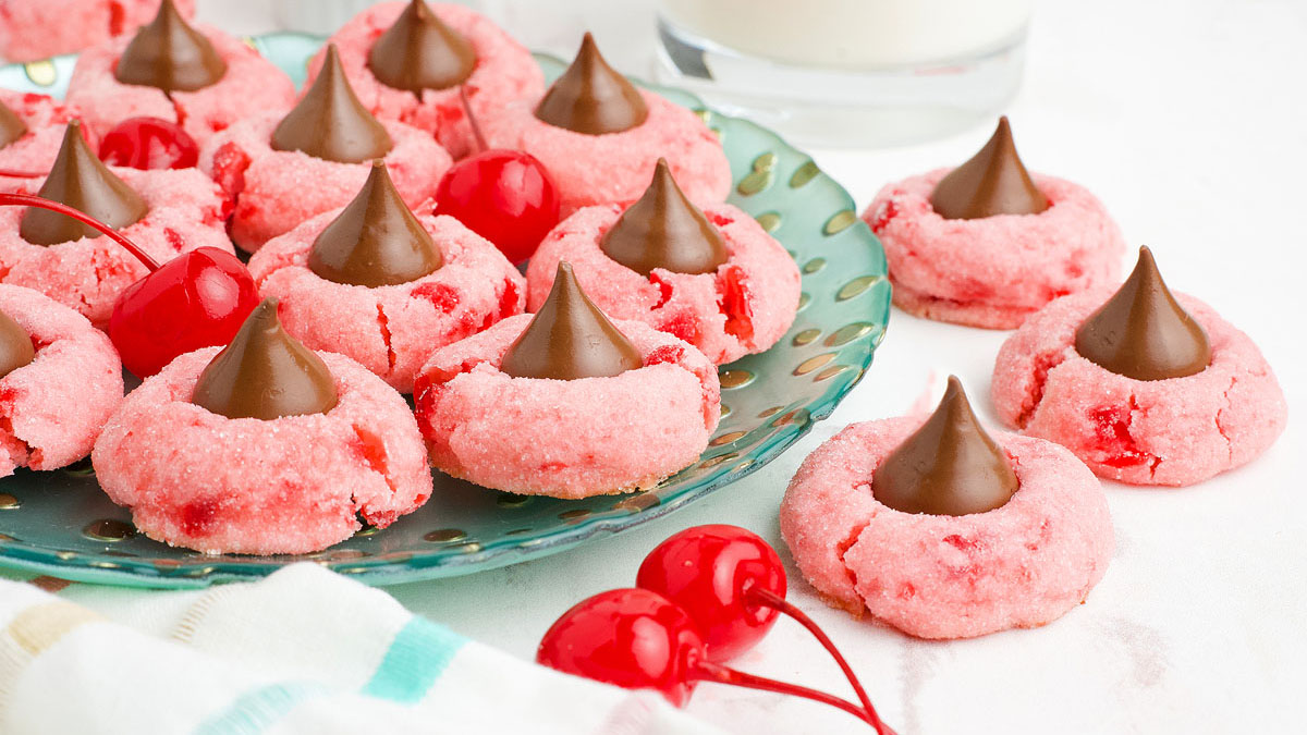 A green plate filled with pink cherry cookies topped with chocolate kisses, surrounded by maraschino cherries and a glass of milk.
