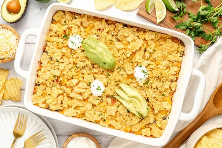 A baked casserole topped with crushed tortilla chips, garnished with avocado slices and dollops of sour cream, surrounded by plates and fresh ingredients on a table.