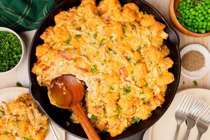 A cast iron skillet filled with cheesy chicken tater tot casserole, garnished with chopped parsley, with a wooden spoon partially serving a portion.