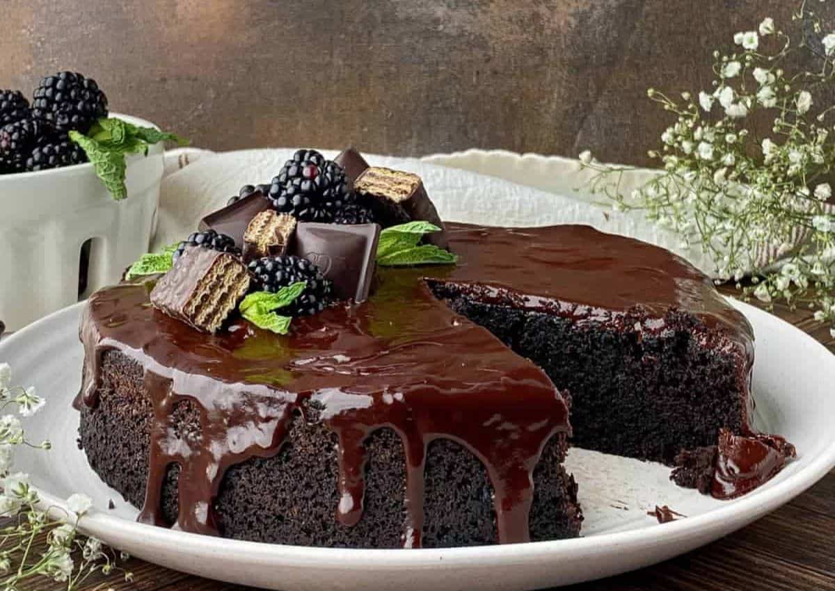 A chocolate cake with glossy chocolate glaze, topped with blackberries, mint leaves, and chocolate wafer pieces, displayed on a white plate with a slice removed.