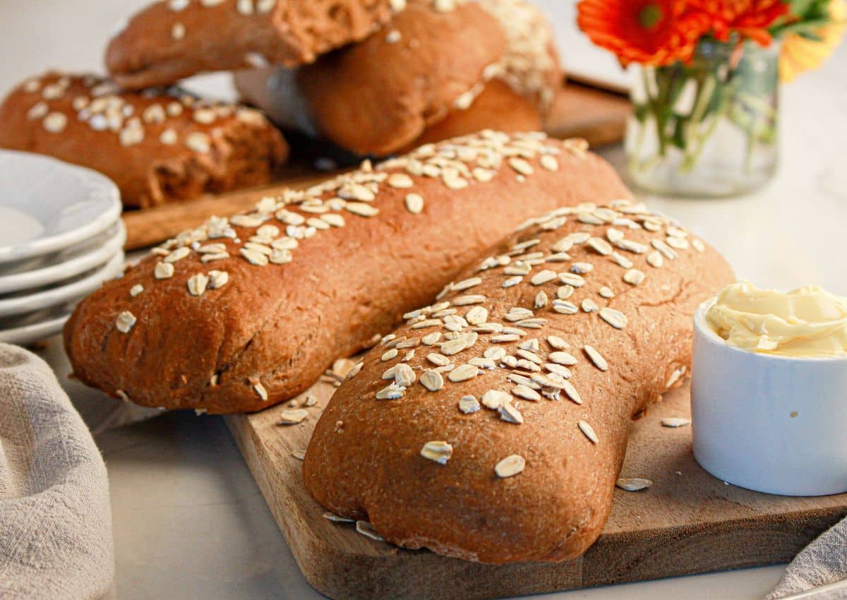 Two loaves of oat-topped bread rest on a wooden board next to a small dish of butter, with plates, a towel, and a vase of flowers in the background.
