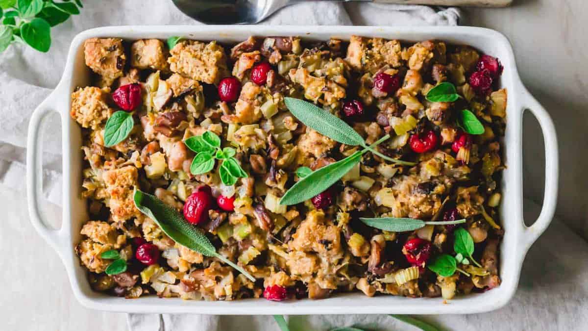 A rectangular white baking dish filled with stuffing topped with cranberries and fresh herbs, including sage and oregano.