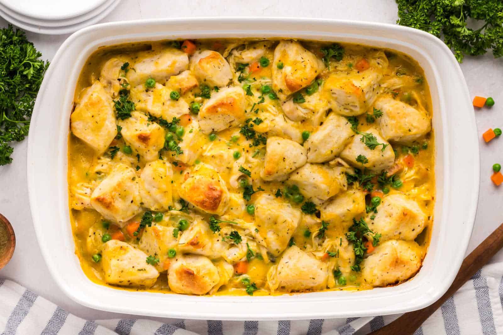 A baked casserole dish filled with biscuit pieces, shredded chicken, peas, carrots, and a creamy yellow sauce, garnished with chopped parsley.